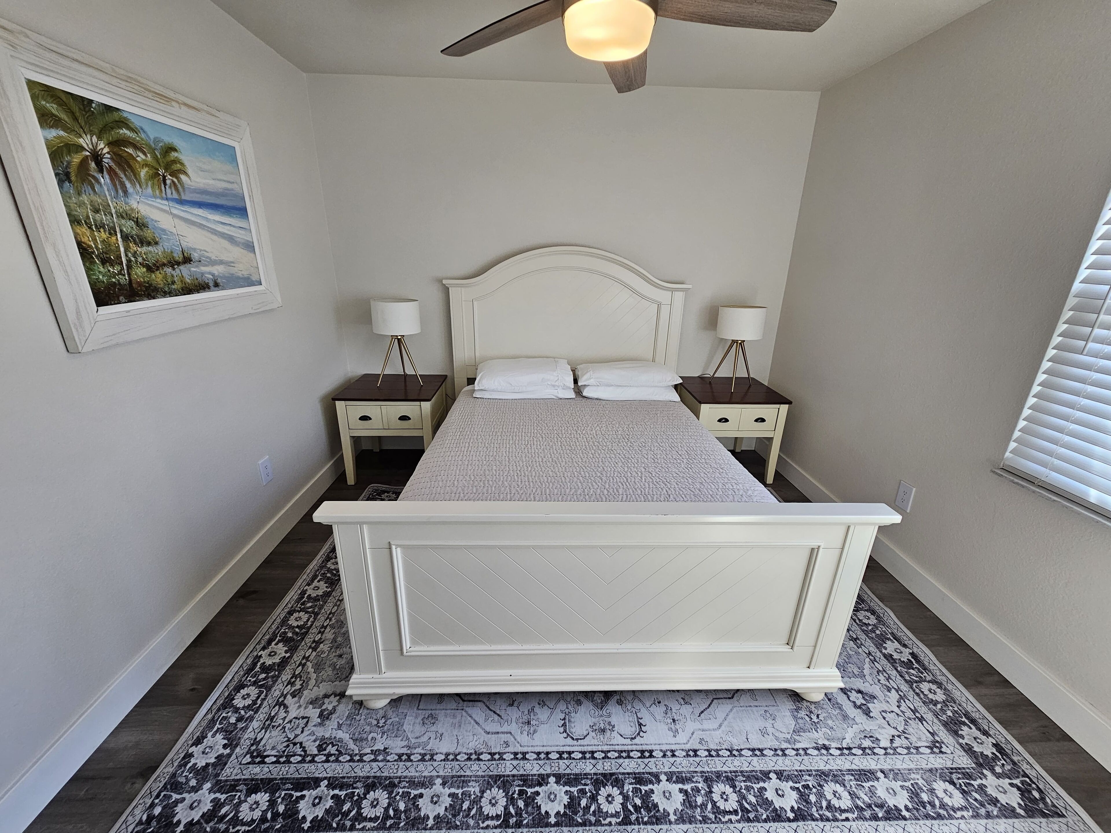 A bedroom with a white bed, two bedside tables with lamps, a ceiling fan, a patterned rug, and a framed picture on the wall.