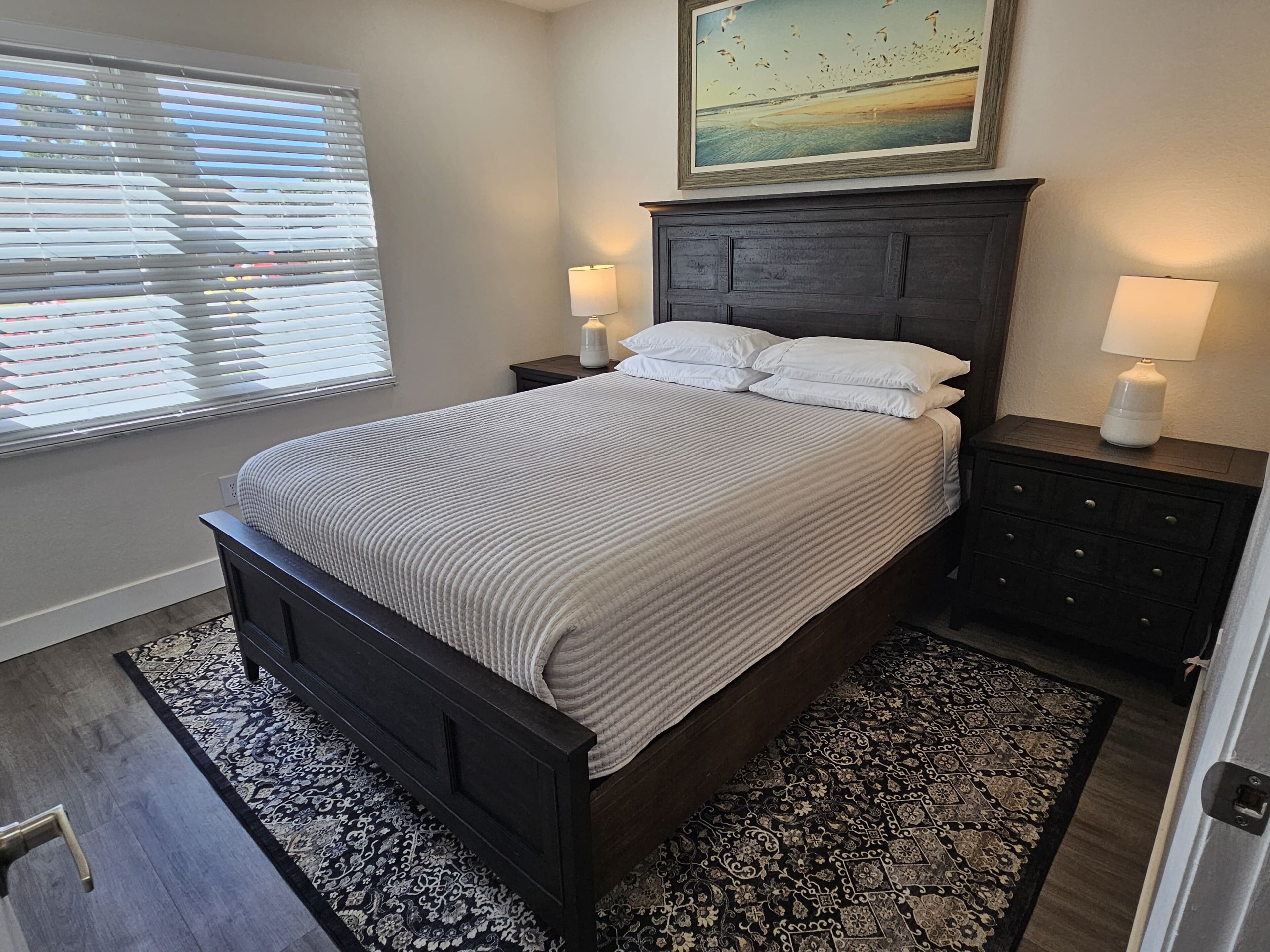 A bedroom with a dark wooden bed, white bedding, two bedside tables with lamps, a patterned rug, and a painting above the bed.