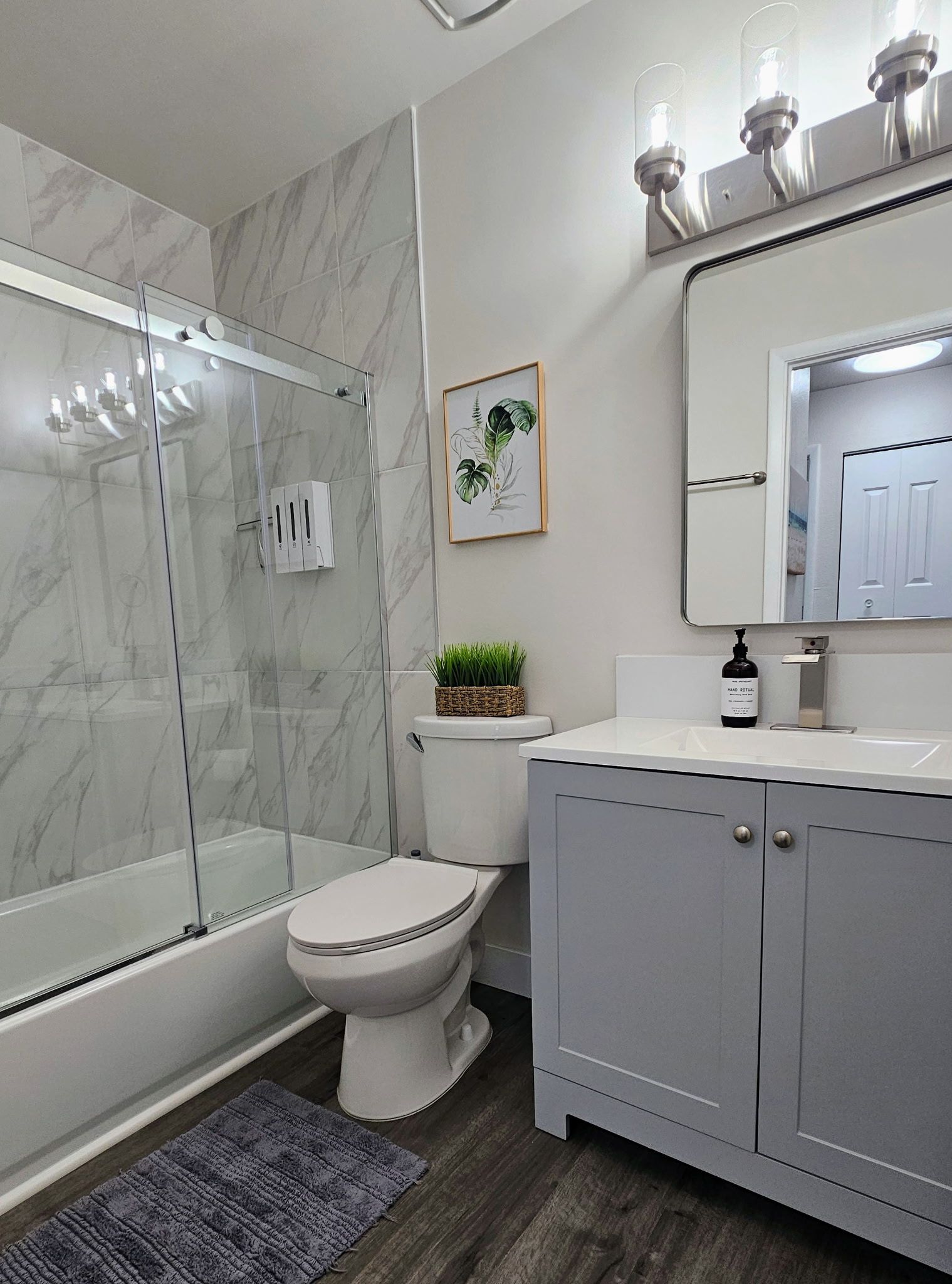 Et badeværelse med brusekabine i glas, toilet, vask med spejl og et botanisk tryk i ramme på væggen.