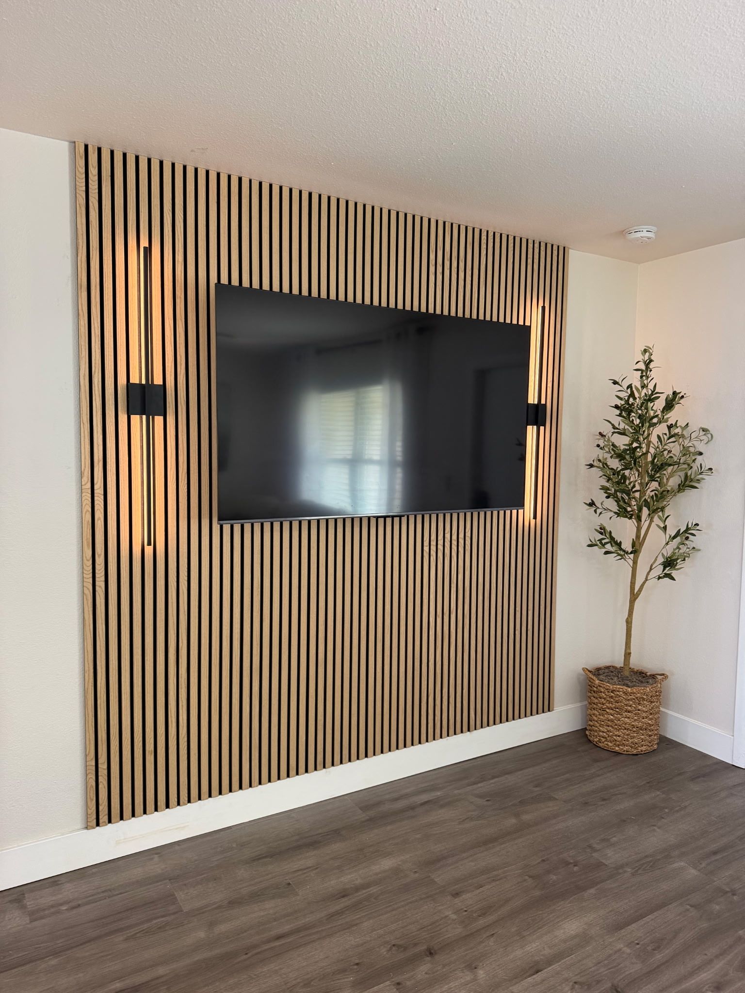 Un soggiorno moderno con una grande TV a schermo piatto montata su una parete di listelli di legno, una pianta in vaso e un pavimento in legno scuro.