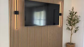A modern living room with a large flat-screen TV mounted on a wooden slat wall, a potted plant, and a dark wooden floor.