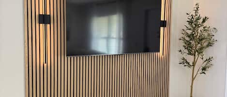 Una sala de estar moderna con un televisor de pantalla plana grande montado en una pared de listones de madera, una planta en maceta y un suelo de madera oscura.