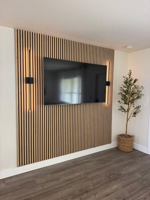 Ein modernes Wohnzimmer mit einem großen Flachbildfernseher an einer Wand aus Holzleisten, einer Topfpflanze und einem dunklen Holzboden.