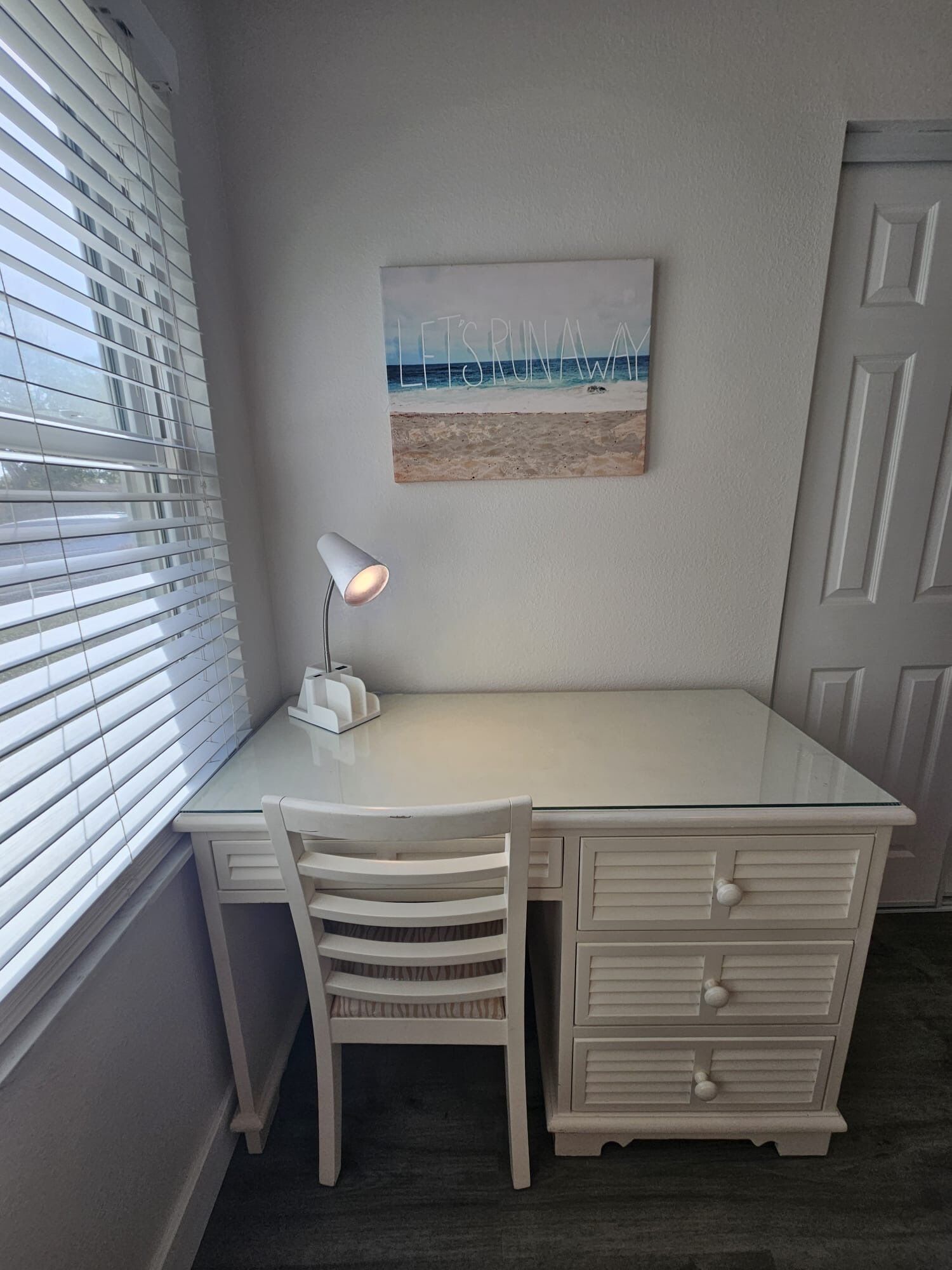 Un bureau blanc avec une lampe, une chaise et un tableau représentant une plage accroché au mur.