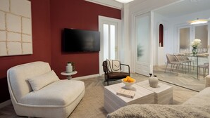 A modern living room with a red accent wall, a flat-screen TV, a comfortable sofa, a coffee table with a bowl of fruit, and a dining area with a glass table and chairs. - Refined & Spacious 3-Bedroom Flat Near Cismigiu, Balcony with Boulevard Views (Bucharest)
