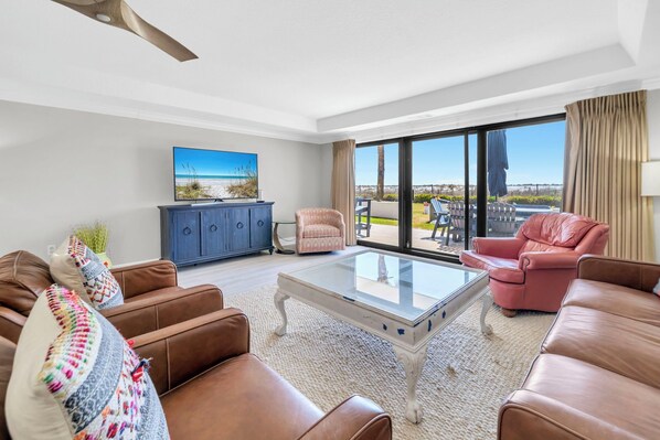 A living room with a glass coffee table, brown leather sofas, a blue cabinet, and a large window with a view of the outdoors.