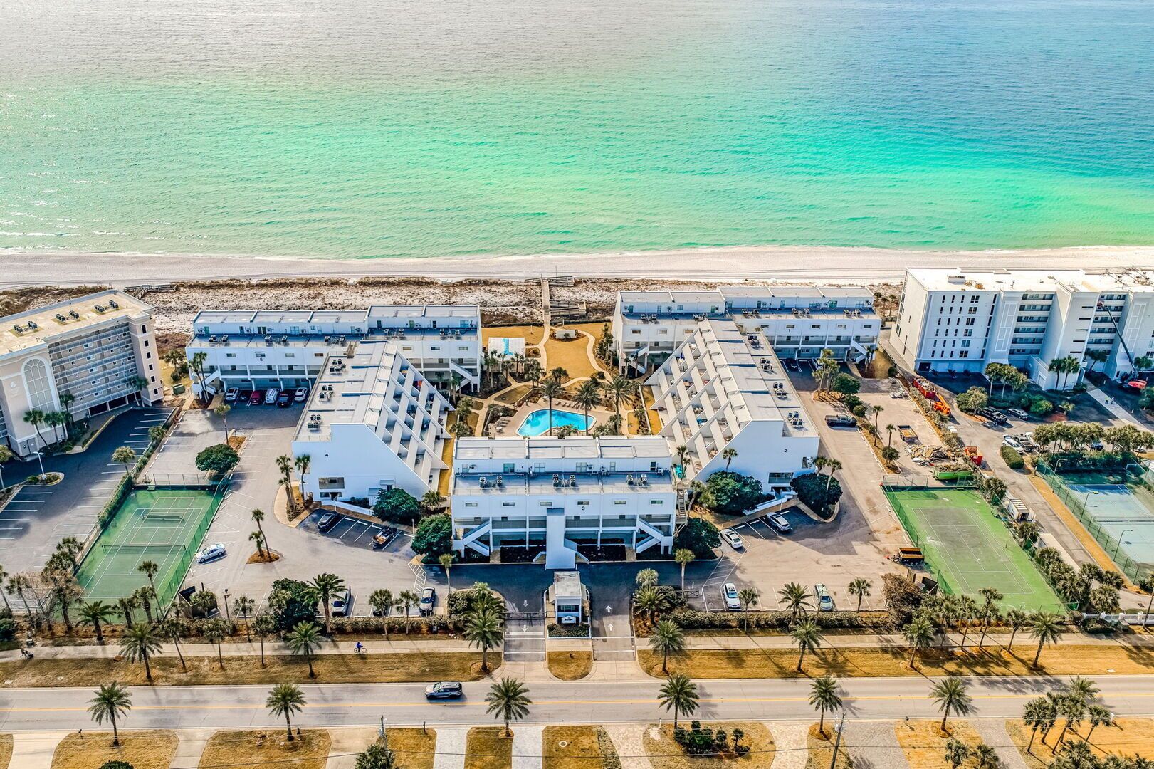 Vista aérea de um hotel à beira-mar, com vários edifícios, uma piscina central e uma praia ao fundo.