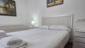 Een netjes opgemaakt bed met witte lakens en kussens, een bedlampje en een nachtkastje met een lamp.