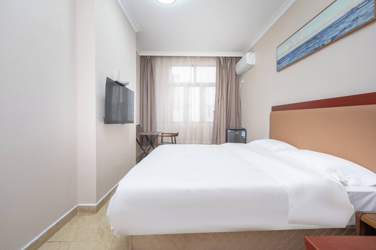 A hotel room with a large bed, a TV mounted on the wall, a small table, and a window with curtains.