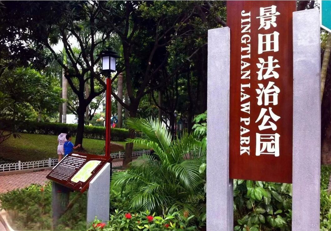 A sign for Jing Tian Law Park with a plaque and a lamp post in the background.