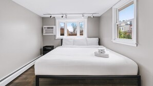 Un dormitorio moderno con una cama grande, una ventana con vista a edificios y un equipo de aire acondicionado en la pared.