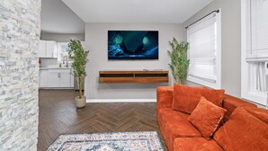 A modern living room with a herringbone floor, a large flat-screen TV, a wooden bench, and a vibrant orange sofa.