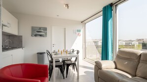 Ein modernes Wohnzimmer mit einer Couch, einem Esstisch mit Stühlen, einer Küchenzeile inklusive Spüle und einem großen Fenster mit Vorhängen.
