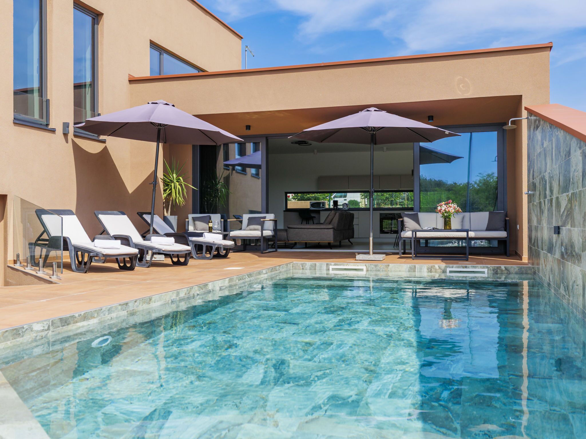 A modern outdoor pool area with lounge chairs and umbrellas, a dining area, and a view of the surrounding landscape.