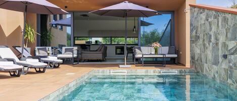 A modern outdoor pool area with lounge chairs and umbrellas, a dining area with a table and chairs, and a stone wall.