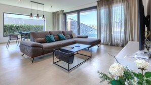 A modern living room with a sectional sofa, a coffee table, and a dining area with a table and chairs. There is a large window with curtains, a ceiling light fixture, and a view of a building outside. - Villa Ana by Interhome (Novigrad (Istra))