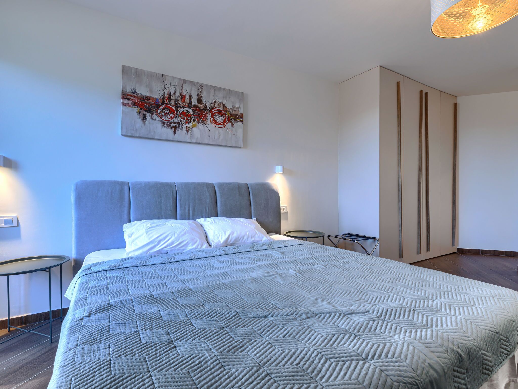 Ein modernes Schlafzimmer mit einem großen Bett, einem grau gepolsterten Kopfteil, einem an der Wand befestigten abstrakten Gemälde und einem Kleiderschrank mit vertikalen Griffen.
