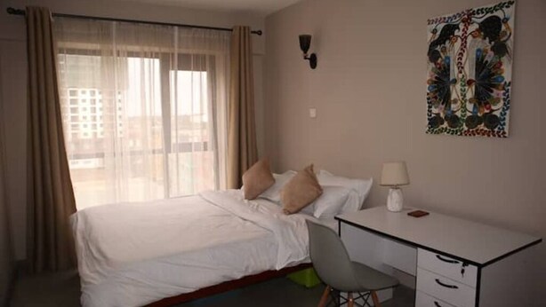 Una habitación de hotel con cama, escritorio y silla. Tiene una ventana con cortinas, una lámpara de pared y una obra de arte.