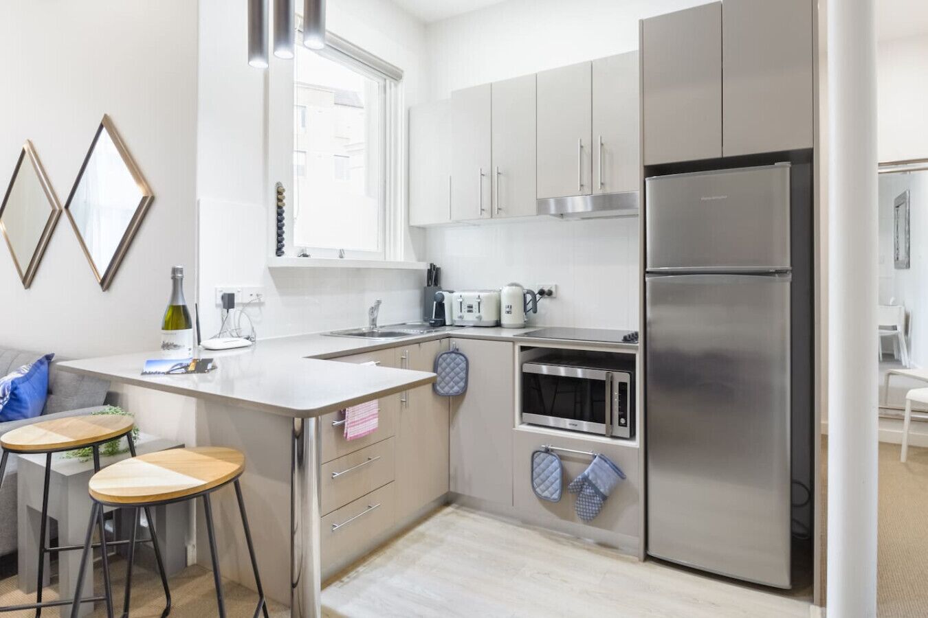 Eine moderne Küche mit Edelstahlgeräten, einem eingebauten Backofen und einem großen Kühlschrank. Zwei Hocker und ein kleiner Tisch mit einer Pflanze stehen dort.