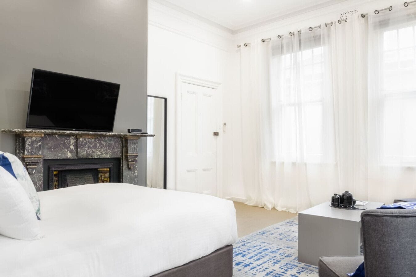 Een hotelkamer met een bed, een televisie boven een open haard, een deur en een raam met dunne gordijnen.