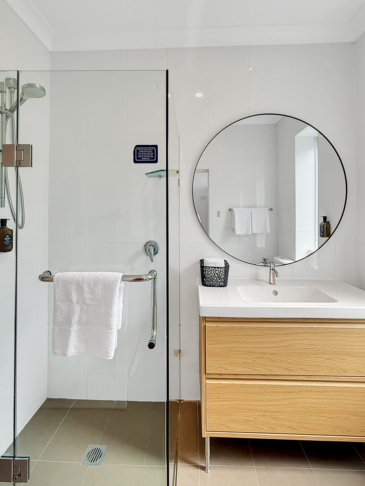 A modern bathroom with a glass shower enclosure, a round mirror, a white sink, and a wooden vanity.