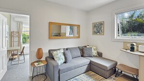 A modern living room with a grey sectional sofa, a small round table, a bench, a large mirror, and a window with a view of palm trees. - Parkside 1 bedroom home, Whisper quiet (North Sydney)