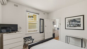 A bedroom with a bed, a dresser, a television, a window, and a kitchen area visible through an open door.