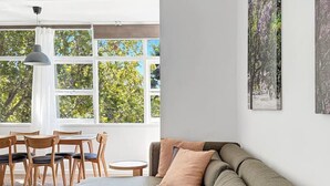 A modern living room with a sectional sofa, a dining area with wooden chairs and a round table, and large windows with a view of trees.