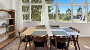 A dining area with a wooden table and chairs, a hanging pendant light, and a view of trees through large windows.