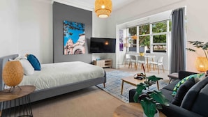 Una habitación de hotel moderna con una cama grande, un televisor, un área de comedor con mesa y sillas, y un ventanal con vistas.