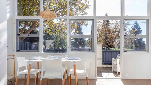 Un comedor luminoso con una mesa dispuesta para cuatro, ventanas grandes y vistas a árboles y una valla.