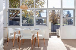 Une salle à manger lumineuse avec une table dressée pour quatre personnes, de grandes fenêtres et une vue sur les arbres et une clôture.