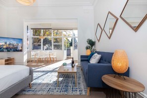 A modern living room with a bed, a blue sofa, a coffee table, and a lamp. There is a large window with a view of trees and a cityscape.