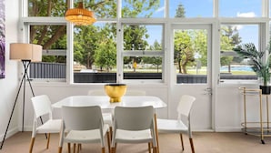 Un comedor con una mesa y sillas blancas, un centro de mesa con un cuenco dorado y ventanas amplias con vistas a árboles y una valla.