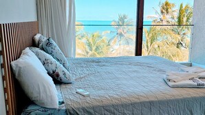 Un dormitorio con una cama grande, cabecera de madera, vistas a palmeras y al mar a través de una ventana, y un aparador blanco.