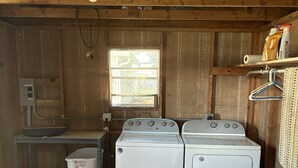 A laundry room with two white Whirlpool washers, a wooden ceiling, a window, and a metal shelf with cleaning supplies. - Charming 2-bedroom, 1-bath lakefront house in the Ocala National Forest (Silver Springs)