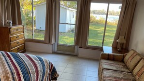 A bedroom with a bed, a dresser, a sofa, and a view of a house and lake through large windows. - Charming 2-bedroom, 1-bath lakefront house in the Ocala National Forest (Silver Springs)