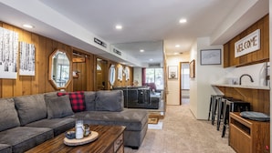 A modern living room with a gray sectional sofa, a wooden coffee table, and a kitchen area with a 'WELCOME' sign.