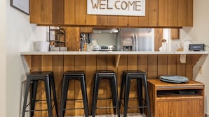 A kitchen with wooden cabinets, a welcome sign, a stove, a refrigerator, and three black stools.