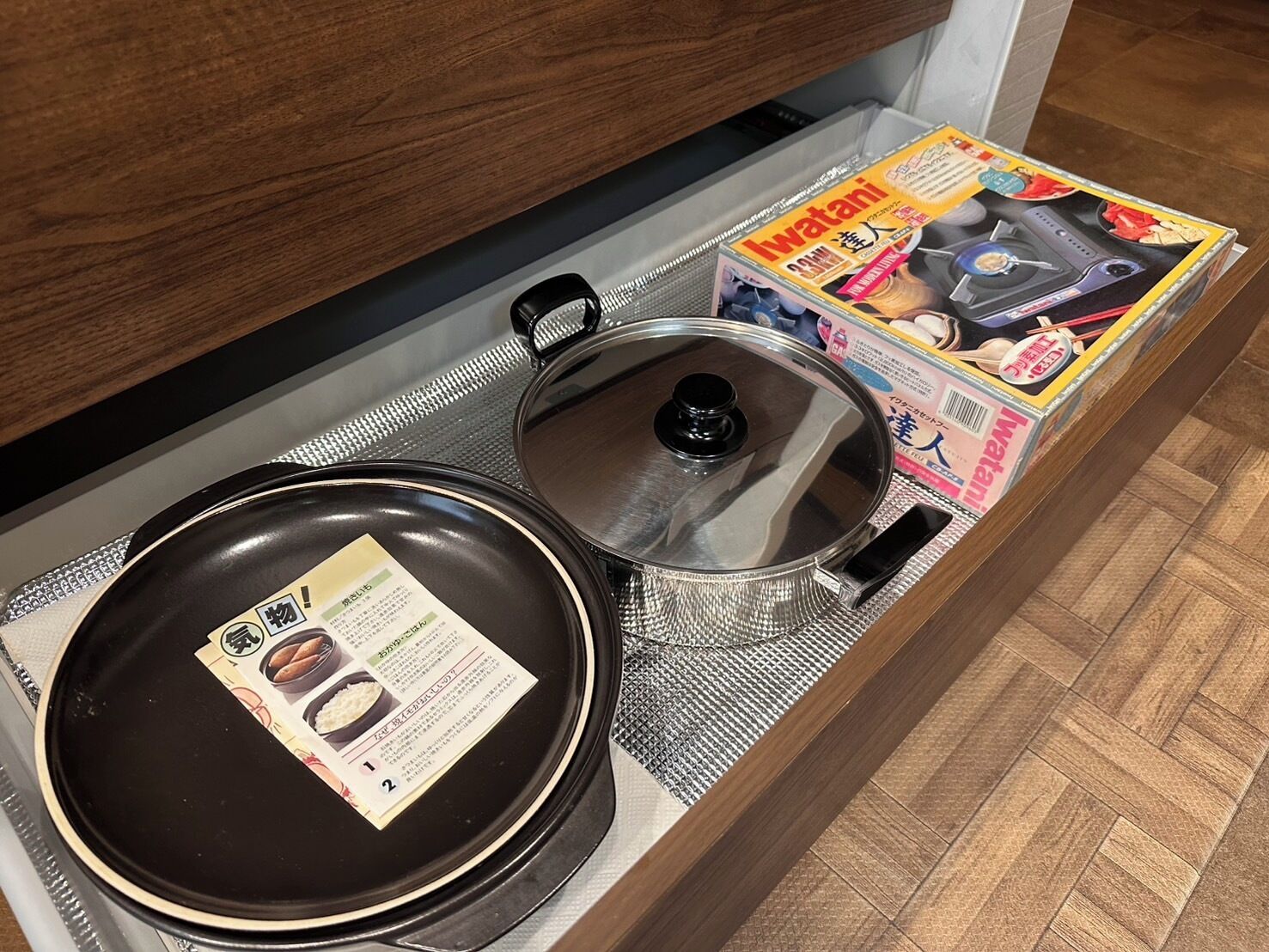 A kitchen drawer with a black frying pan, a stainless steel pot, and a magazine.