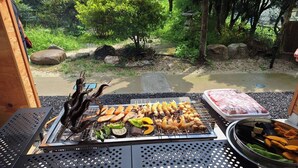 A grill with various meats and vegetables, a table with a metal frame, and a view of a garden with trees and rocks. - Warouan (Nikko)