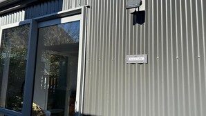 A metal building with a sliding glass door, a sign reading 'Custom Cabin,' and a small black object on the wall. - Custom Cabin -  In Town - Hot Tub - Super King Bed (Queenstown)