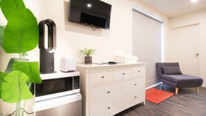 A modern room with a flat-screen TV, a microwave, a white dresser, a grey chair, and a large green plant. - Custom Cabin -  In Town - Hot Tub - Super King Bed (Queenstown)