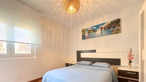 Un dormitorio con cama, mesitas de noche, repisa en la pared, ventana con persianas y un cuadro.