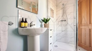 A bathroom with a white sink, two bottles of shampoo, a mirror, a shower, and a wooden door. - Shepherds Rest - Four bedroom home (County Kerry)