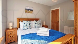 A neatly made bed with a blue and white blanket, wooden headboard, and matching pillows. There is a wooden nightstand with a lamp and a small drawer. A framed picture hangs on the wall above the bed. - Shepherds Rest - Four bedroom home (County Kerry)
