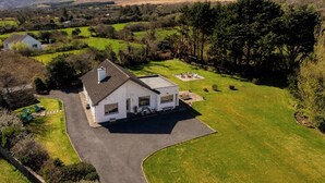 A modern house with a large driveway and a well-maintained lawn, surrounded by a rural landscape with mountains in the background. - Owenmore Lodge 3 bedroom home (County Kerry)