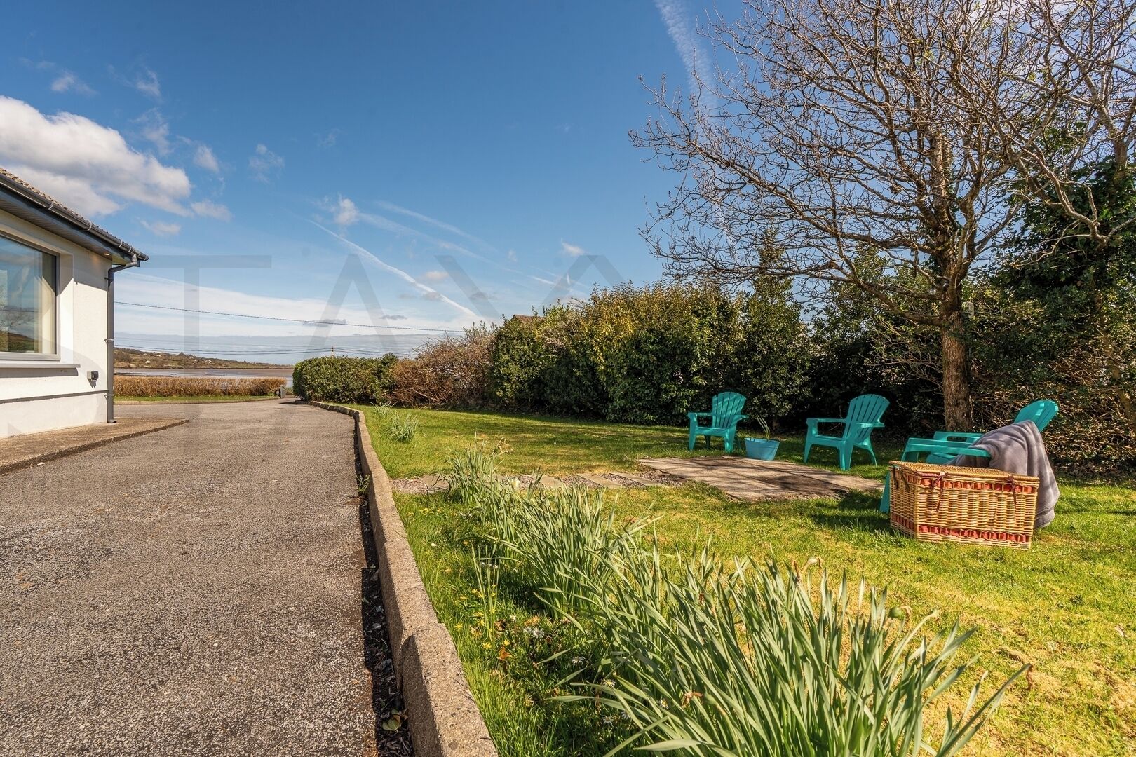 A residential property with a paved driveway leading to a house, a grassy yard with a tree, and outdoor seating with chairs and a basket.