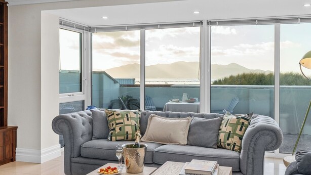 A modern living room with a grey sofa, a coffee table with a book and a plate of food, and a large window offering a view of the ocean and mountains.