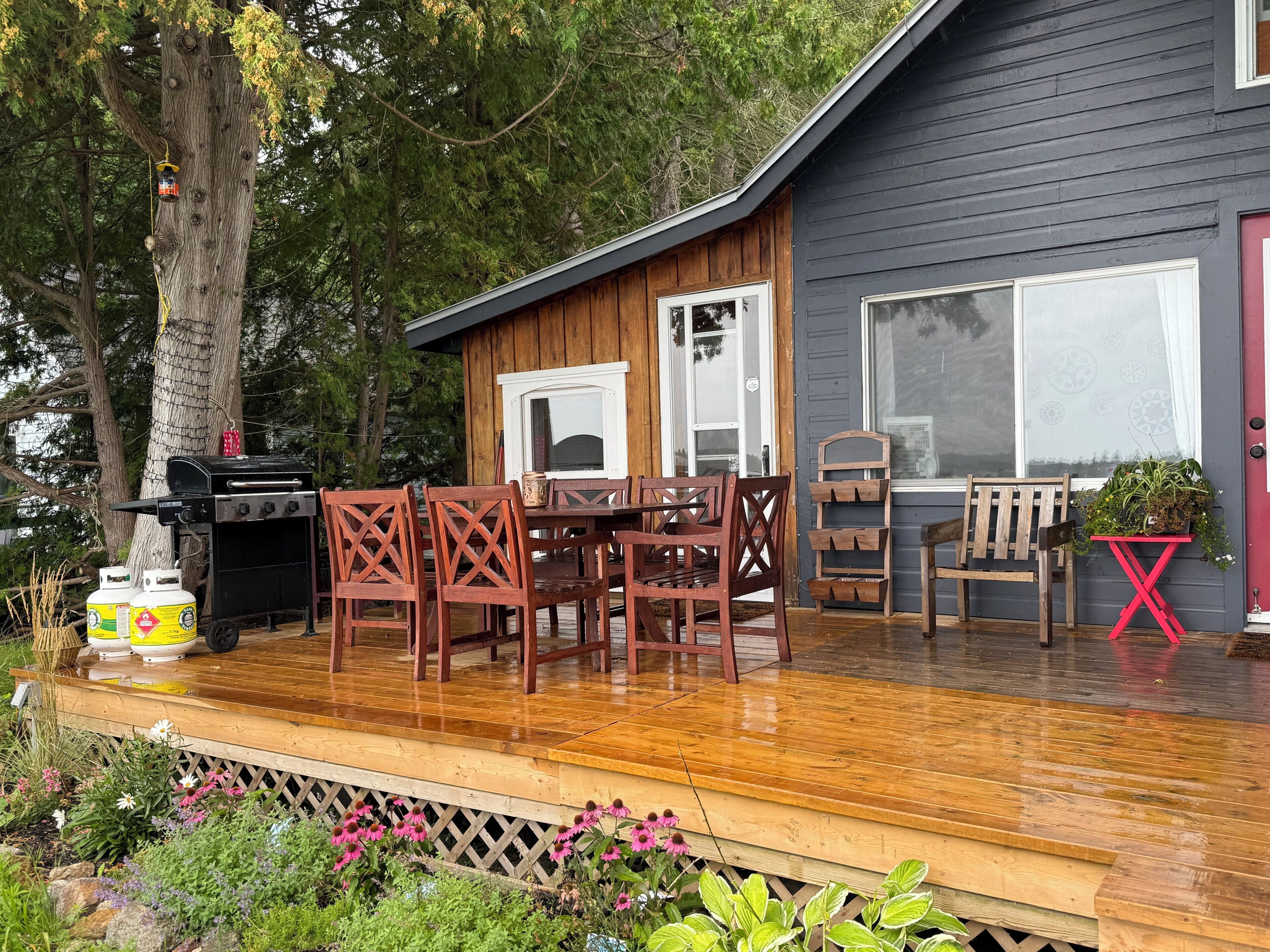 A wooden deck with a grill, dining table, and chairs, a small table with plants, and a red chair.